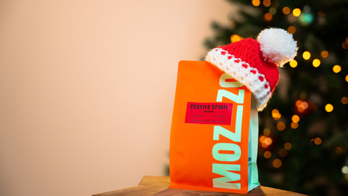 a bag of festive spirit coffee beans with a red & white Santa hat, Christmas tree in background