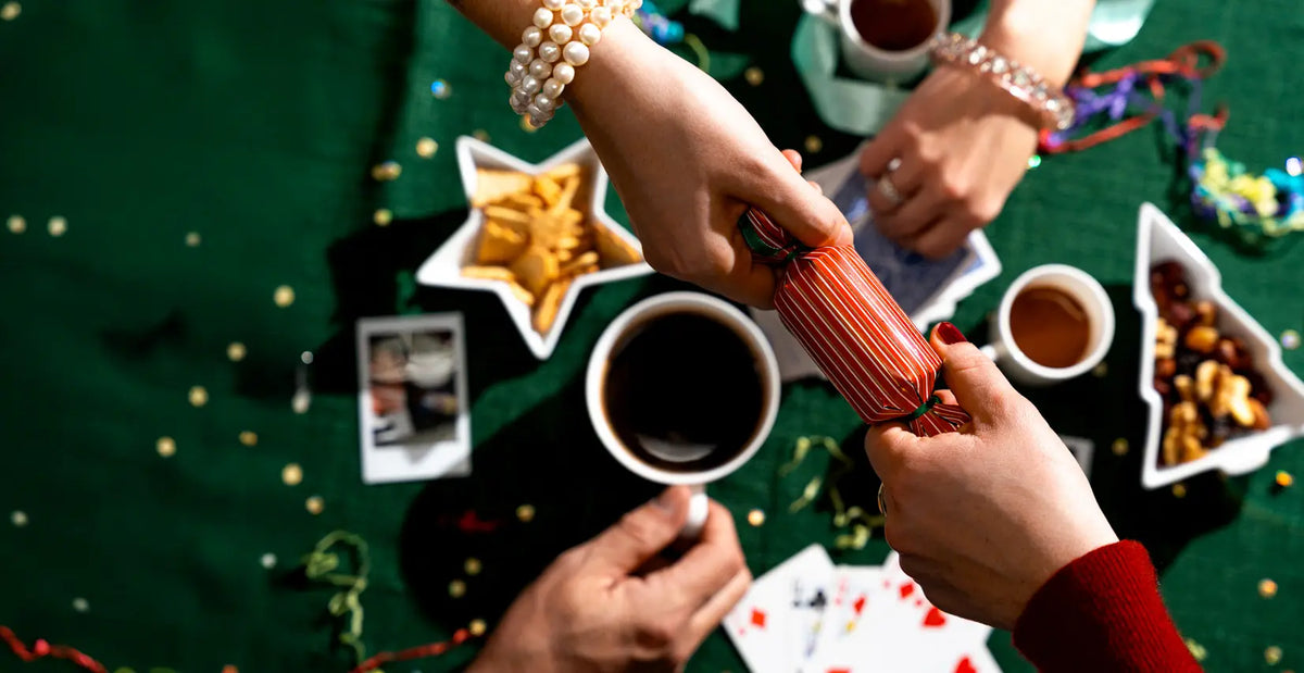 Two hands pulling a Christmas cracker above a Christmas table with coffees and playing cards