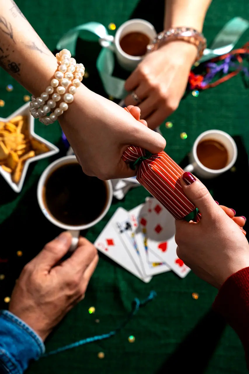 Two hands pulling a Christmas cracker above a Christmas table with coffees and playing cards